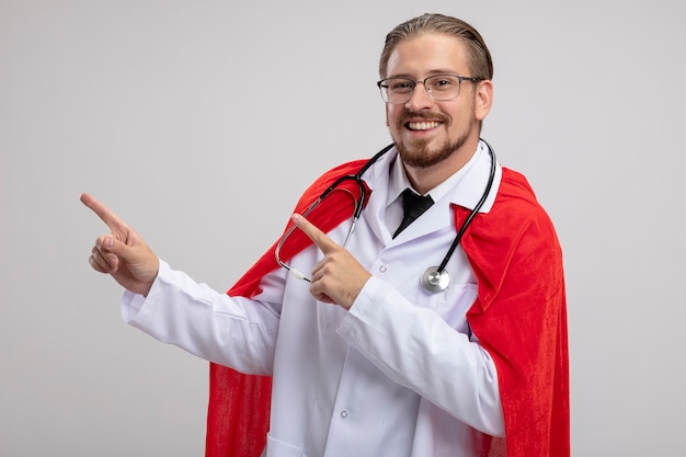 Kostenloses Foto lächelnder junger superheldenmann, der medizinisches gewand mit stethoskop und brille zeigt, zeigt seitlich lokalisiert auf weißem hintergrund mit kopienraum