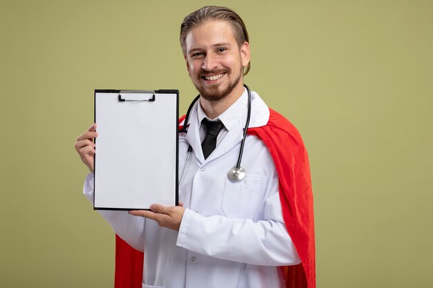 Lächelnder junger Superheldenmann, der Stethoskop mit medizinischem Gewand trägt, das Klemmbrett lokalisiert auf olivgrünem Hintergrund hält