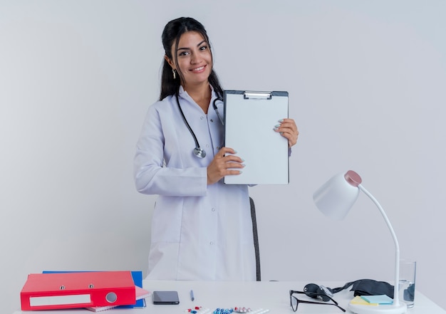 Lächelnder junger weiblicher Arzt, der medizinisches Gewand und Stethoskop trägt, sitzt am Schreibtisch mit medizinischen Werkzeugen, die Klemmbrett isoliert zeigen