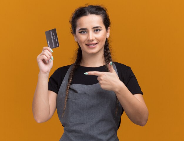 Lächelnder junger weiblicher Friseur im einheitlichen Halten und zeigt auf Kreditkarte lokalisiert auf orange Wand