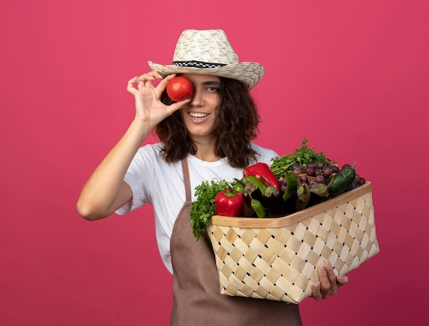 Lächelnder junger weiblicher Gärtner in der Uniform, die Gartenhut hält, der Gemüsekorb hält, der Blickgeste mit Tomate lokalisiert auf Rosa zeigt