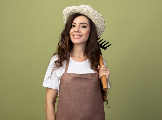 Lächelnder junger weiblicher Gärtner in der Uniform, die Gartenhut trägt, hält Rechen auf Schulter lokalisiert auf olivgrüner Wand