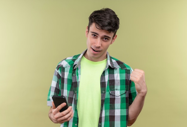 Lächelnder kaukasischer junger Mann, der grünes Hemd hält, das Telefon zeigt, das ja Geste auf lokalisiertem grünem Hintergrund zeigt