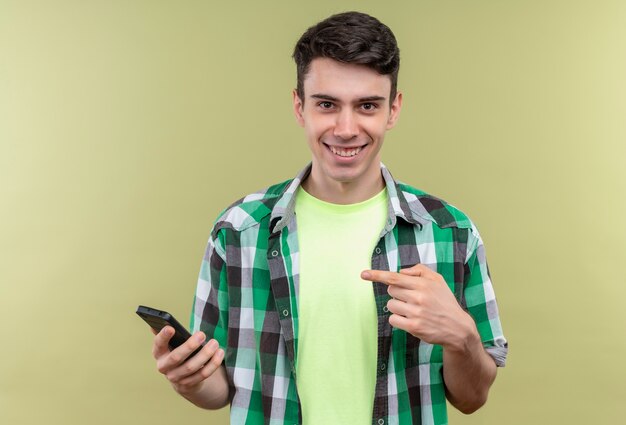 Lächelnder kaukasischer junger Mann, der grünes Hemd trägt, zeigt auf Telefon in seiner Hand auf lokalisiertem grünem Hintergrund
