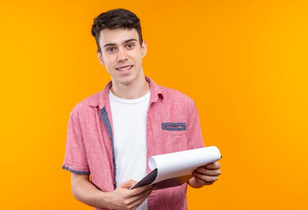 Lächelnder kaukasischer junger Mann, der rosa Hemd hält, das Klemmbrett auf lokalisiertem orange Hintergrund hält