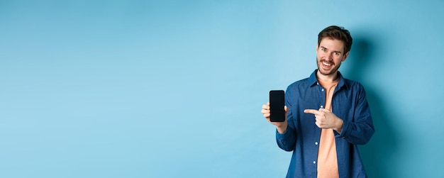Lächelnder kaukasischer Mann, der auf einem leeren Smartphone-Bildschirm zeigt, auf das Handy zeigt und zufrieden aussieht
