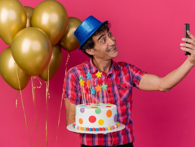Lächelnder kaukasischer Parteimann mittleren Alters, der Parteihut trägt, der nahe Luftballons hält, die Geburtstagstorte halten, die selfie lokalisiert auf purpurrotem Hintergrund nimmt