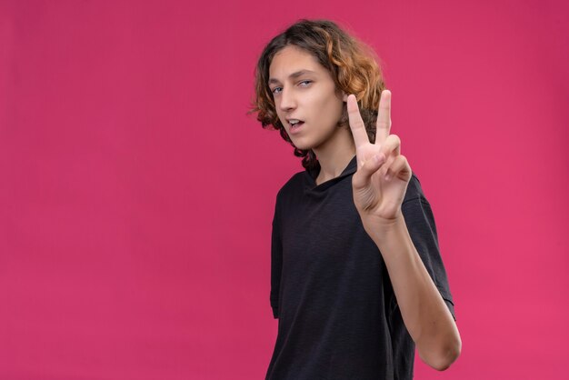 Lächelnder Kerl mit langen Haaren im schwarzen T-Shirt, das eine viva Gasture auf rosa Wand zeigt