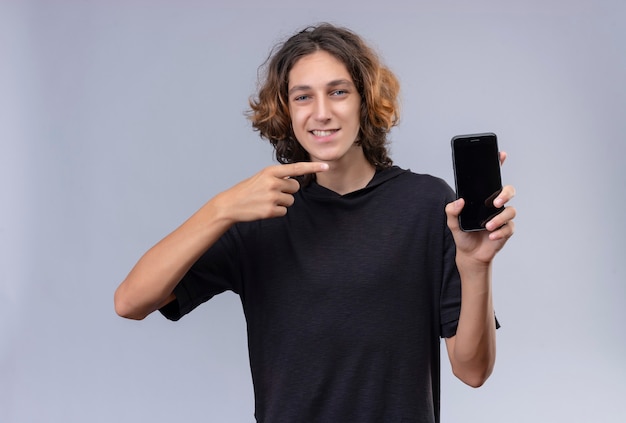 Lächelnder Kerl mit langen Haaren im schwarzen T-Shirt, der ein Telefon hält und auf Telefon auf weißer Wand zeigt