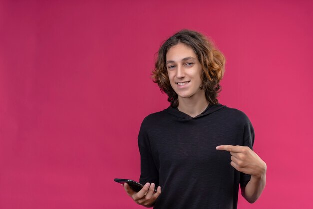 Lächelnder Kerl mit langen Haaren im schwarzen T-Shirt, der ein Telefon hält und zur Seite auf rosa Wand zeigt