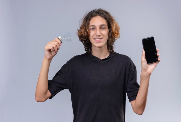 Lächelnder Kerl mit langen Haaren im schwarzen T-Shirt, der ein Telefon und eine Bankkarte auf weißer Wand hält