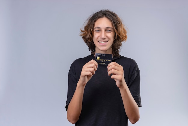 Kostenloses Foto lächelnder kerl mit langen haaren im schwarzen t-shirt, der eine bankkarte auf weißer wand hält