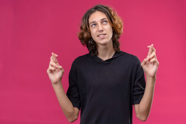 Lächelnder Kerl mit langen Haaren im schwarzen T-Shirt, der seine Finger auf rosa Wand kreuzt