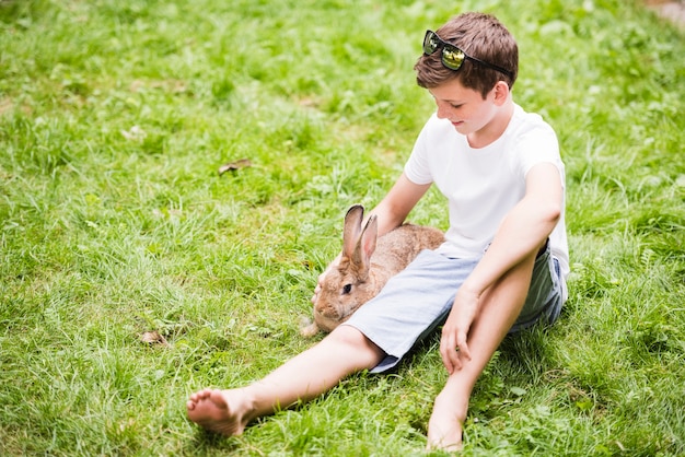 Lächelnder kleiner junge, der mit kaninchen auf grünem gras sitzt