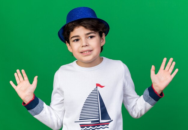 Lächelnder kleiner Junge mit blauem Partyhut, der die Hände isoliert auf grüner Wand ausbreitet
