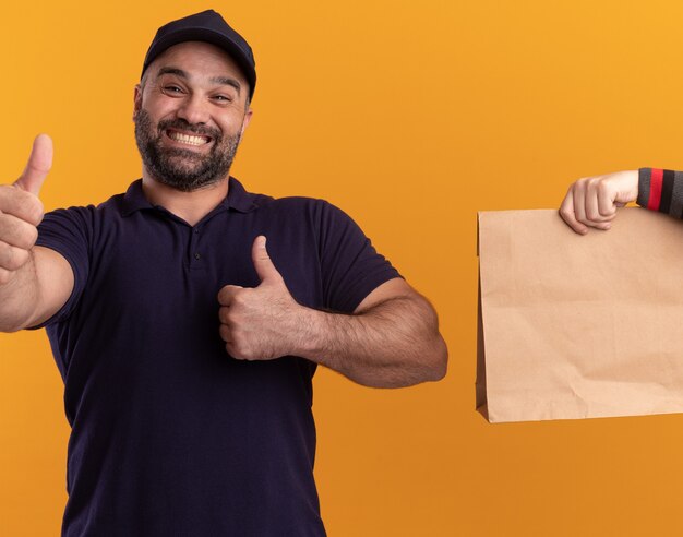 Lächelnder Lieferbote mittleren Alters in Uniform und Mütze, die dem Kunden Papiernahrungsmittelpaket geben, das Daumen oben isoliert auf gelber Wand zeigt