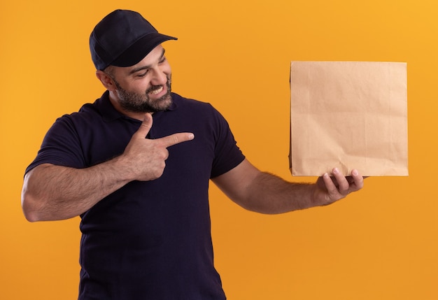 Lächelnder Lieferbote mittleren Alters in Uniform und Mützenhaltung und Punkte auf Papiernahrungsmittelverpackung lokalisiert auf gelber Wand