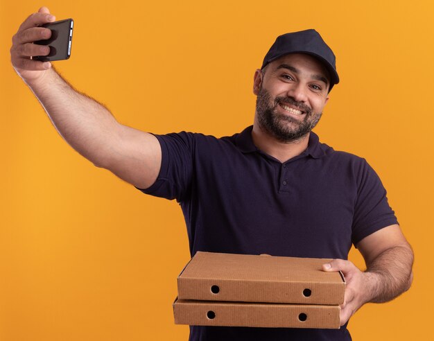 Lächelnder Liefermann mittleren Alters in Uniform und Mütze mit Pizzakartons und macht ein Selfie isoliert auf gelber Wand