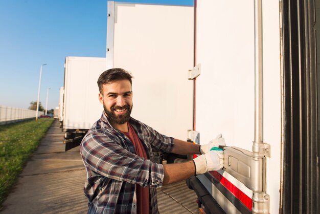 Lächelnder LKW-Fahrer in Arbeitshandschuhen, die die Hintertüren des LKW-Anhängers öffnen oder schließen und Waren auf Transport prüfen