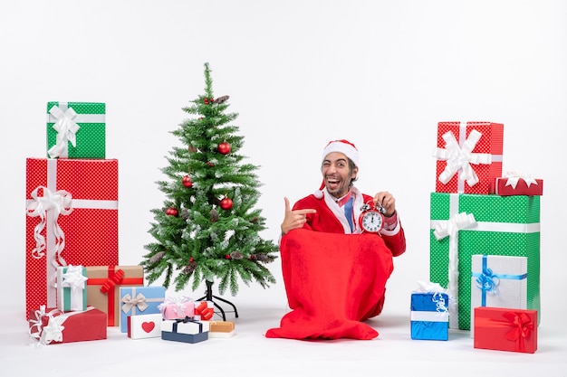 Kostenloses Foto lächelnder lustiger junger mann feiern neujahrs- oder weihnachtsfeiertag, der auf dem boden sitzt und uhr nahe geschenken und geschmücktem weihnachtsbaum hält