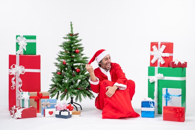 Lächelnder lustiger junger Mann verkleidet als Weihnachtsmann mit Geschenken und geschmücktem Weihnachtsbaum, der auf dem Boden auf weißem Hintergrund sitzt