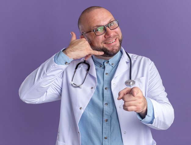 Lächelnder männlicher Arzt mittleren Alters, der ein medizinisches Gewand und ein Stethoskop mit einer Brille trägt und darauf zeigt, die Anrufgeste isoliert auf lila Wand zu tun?