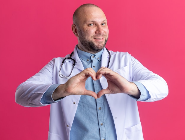 Kostenloses Foto lächelnder männlicher arzt mittleren alters, der ein medizinisches gewand und ein stethoskop trägt und ein herzzeichen isoliert auf einer rosa wand macht