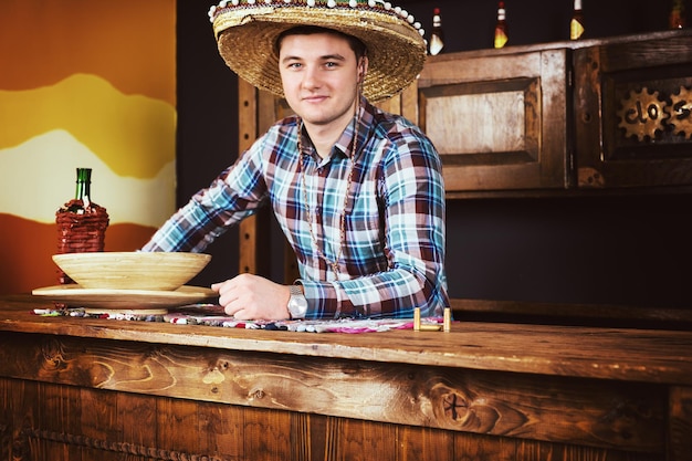 Lächelnder männlicher barkeeper in einem sombrero lehnte sich an die bartheke in einem mexikanischen pub, ein regal mit würzigen saucen im hintergrund