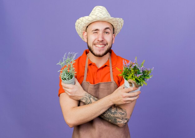 Lächelnder männlicher gärtner, der gartenhut trägt, blinzelt auge und verschränkt arme, die blumentöpfe halten