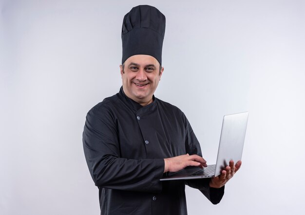 Lächelnder männlicher Koch mittleren Alters in Kochuniform benutzte Laptop in seiner Hand