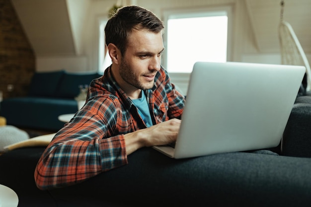 Lächelnder Mann, der auf dem Laptop im Internet surft, während er sich zu Hause auf dem Sofa entspannt