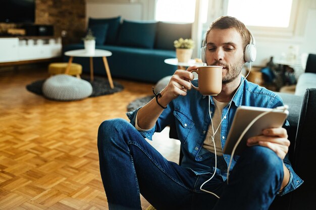 Lächelnder Mann, der auf einem digitalen Tablet im Internet surft, während er sich zu Hause entspannt und Kaffee trinkt