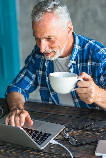 Lächelnder Mann, der Kaffeetasse unter Verwendung des Laptops über dem hölzernen Schreibtisch hält