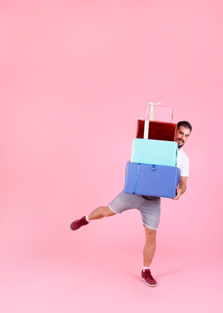 Lächelnder Mann, der mit Stapel bunten Geschenkboxen gegen rosa Hintergrund balanciert