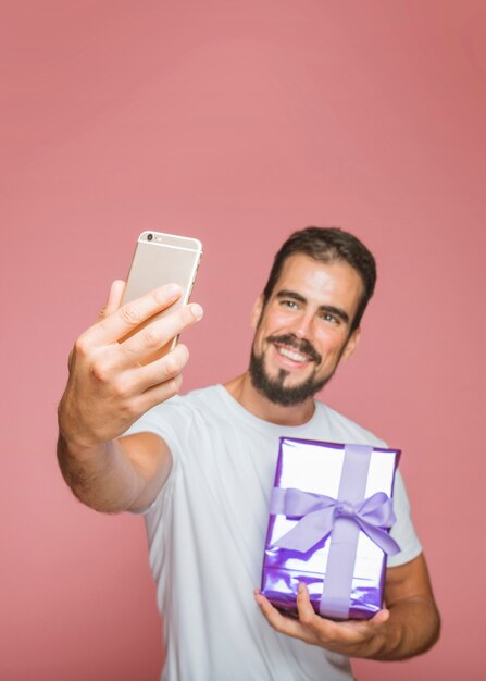 Lächelnder Mann, der purpurrote Geschenkbox selfie vom Mobiltelefon nehmend hält