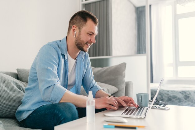 lächelnder Mann im Hemd sitzt entspannt auf Sofa zu Hause am Tisch und arbeitet online am Laptop