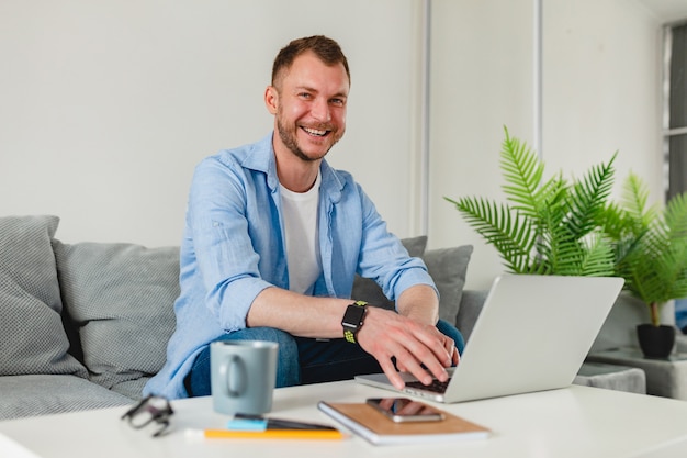 lächelnder Mann im Hemd sitzt entspannt auf Sofa zu Hause am Tisch und arbeitet online auf Laptop von zu Hause aus