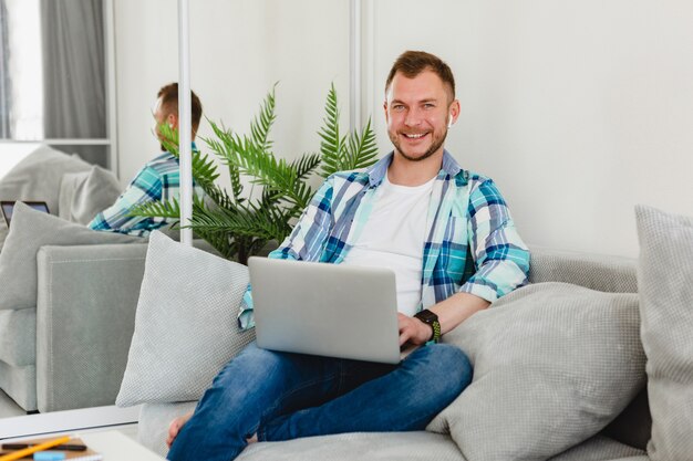 lächelnder Mann im Hemd sitzt entspannt auf Sofa zu Hause am Tisch und arbeitet online auf Laptop von zu Hause aus