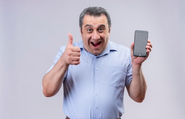 Lächelnder Mann mittleren Alters, der blaues Hemd hält, das Handy hält und Daumen oben zeigt, während auf einem weißen Hintergrund stehend
