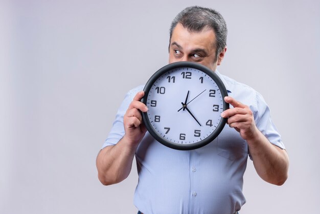 Lächelnder Mann mittleren Alters, der blaues vertikales gestreiftes Hemd hält, das Wanduhr zeigt Zeit zeigt, während auf einem weißen Hintergrund stehend