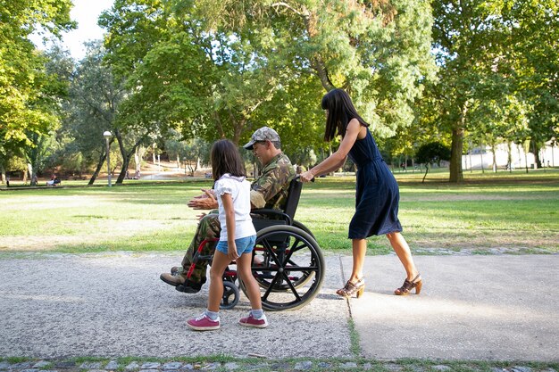 Lächelnder Militärmann, der mit Familie im Stadtpark geht. Langhaarige Mutter schiebt Rollstuhl. Kleines Mädchen, das mit behindertem Vater geht und spricht. Familien-Outdoor-, Wochenend- und Behinderungskonzept