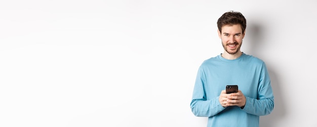 Kostenloses Foto lächelnder moderner mann, der smartphone benutzt und kameramann im sweatshirt mit handy erfreut anschaut
