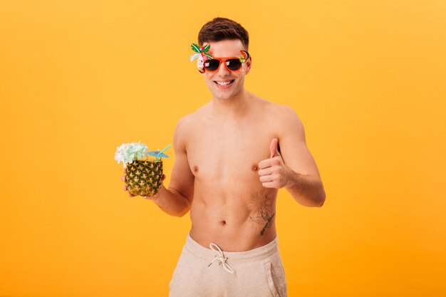 Lächelnder nackter Mann in den kurzen Hosen und in der ungewöhnlichen Sonnenbrille, die Cocktail halten, während Daumen zeigen