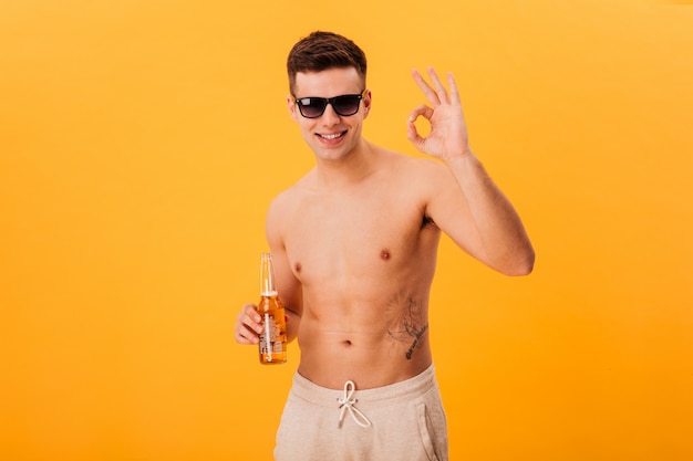 Lächelnder nackter Mann in Shorts und Sonnenbrille hält Flasche Bier und zeigt ok Zeichen über gelb