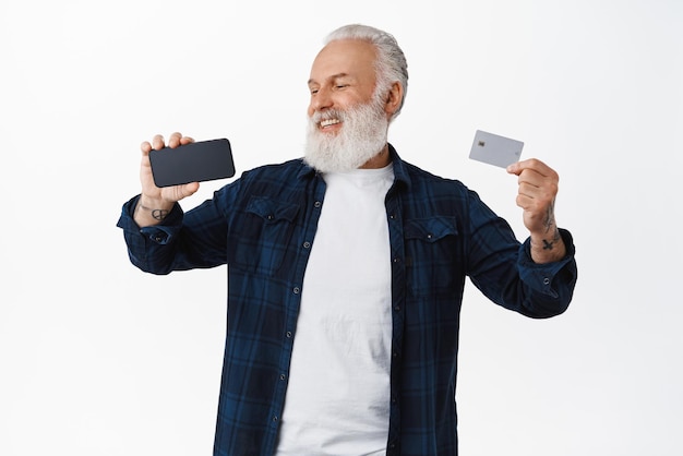 Lächelnder reifer Mann, der die Kreditkarte der Copyspace-Bank zeigt und zufrieden und glücklich auf den horizontalen Stand des Smartphone-Bildschirms vor weißem Hintergrund schaut