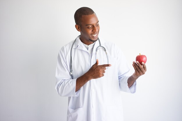 Lächelnder schwarzer männlicher Doktor, der auf roten Apfel hält und zeigt.