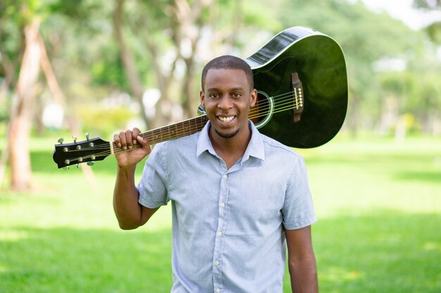 Lächelnder schwarzer Mann, der Gitarre auf Schulter im Park hält