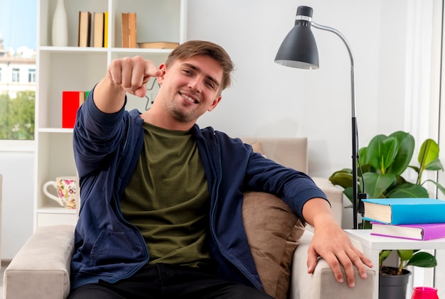 Lächelnder selbstbewusster junger blonder gutaussehender Mann sitzt auf Sessel und zeigt auf Kamera im Wohnzimmer