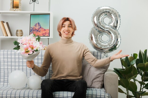 Lächelnder, sich ausbreitender, gutaussehender Kerl am glücklichen Frauentag, der einen Blumenstrauß auf dem Sofa im Wohnzimmer hält