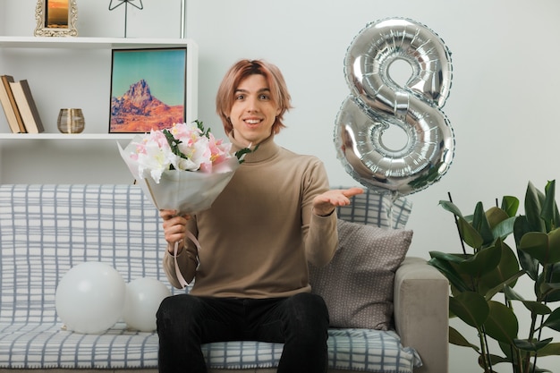 Lächelnder, sich ausbreitender, gutaussehender Kerl am glücklichen Frauentag, der einen Blumenstrauß auf dem Sofa im Wohnzimmer hält